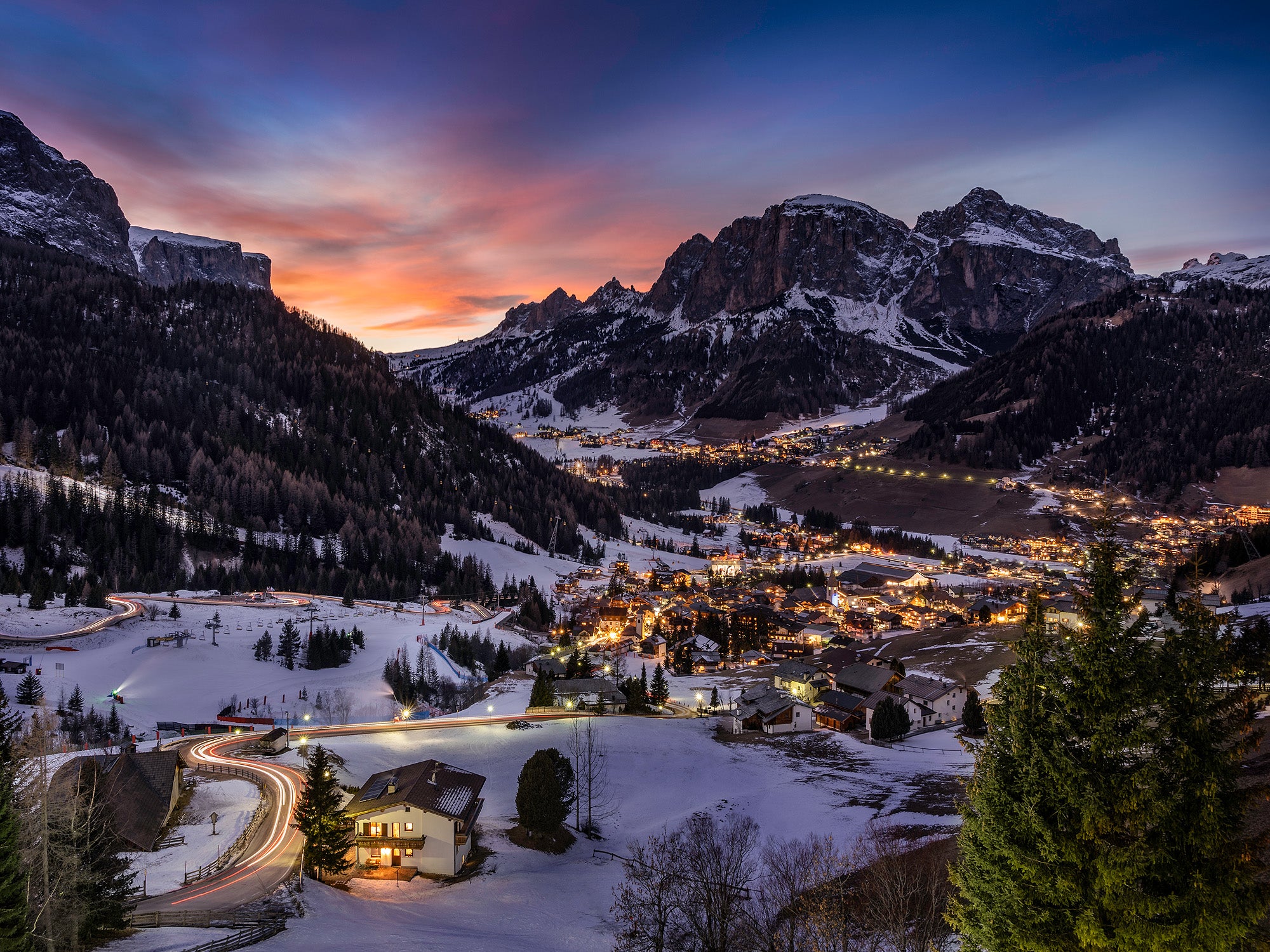 Snow-covered Pyrenean mountain village in Andorra at dusk with winding road and colorful sky in the background.
