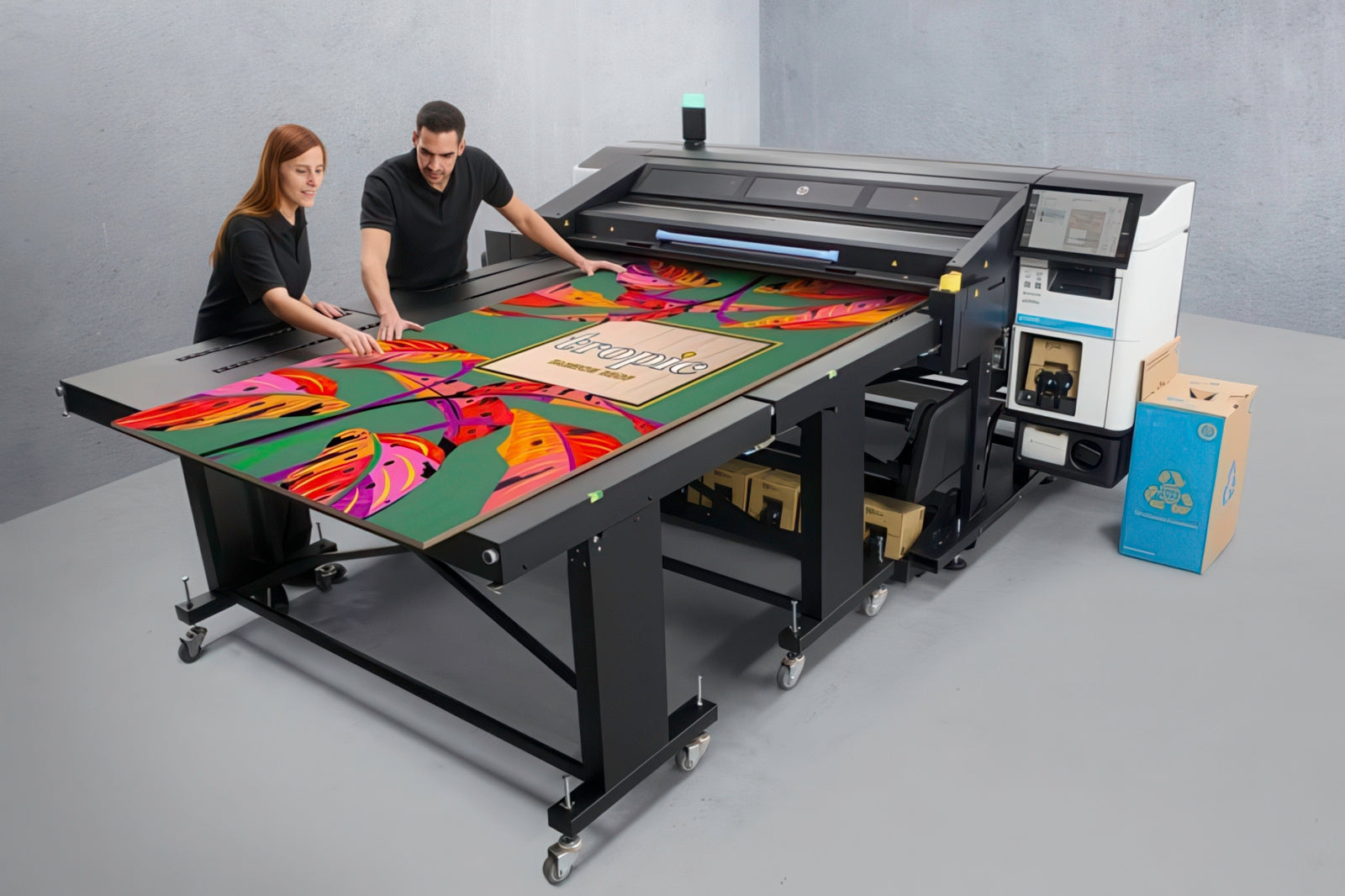 Two people operating a large flatbed printer creating colorful printed artwork on a flat surface.